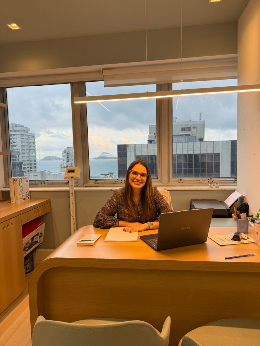 Dra. Gabriela Diogo à mesa do consultório de alergia em Ipanema, com o mar e as Ilhas Cagarras ao fundo